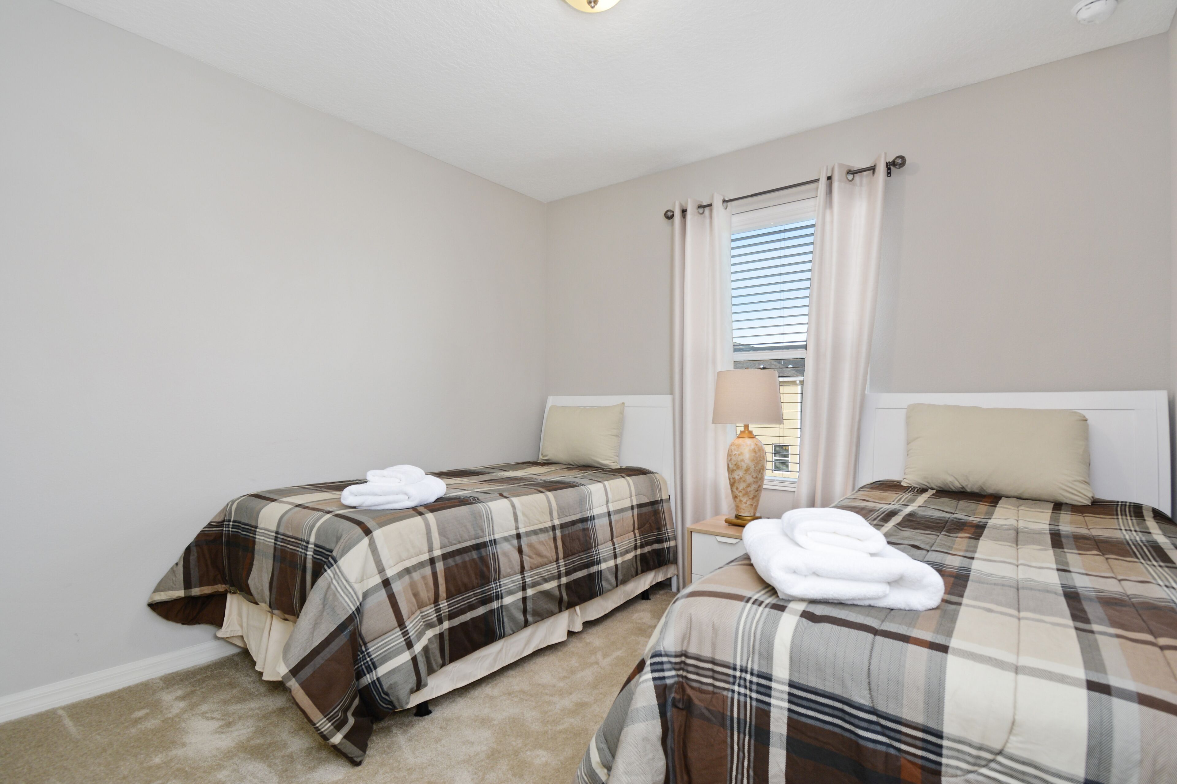 A bedroom with two beds, each with a plaid bedspread and white pillows. There is a nightstand with a lamp between the beds.