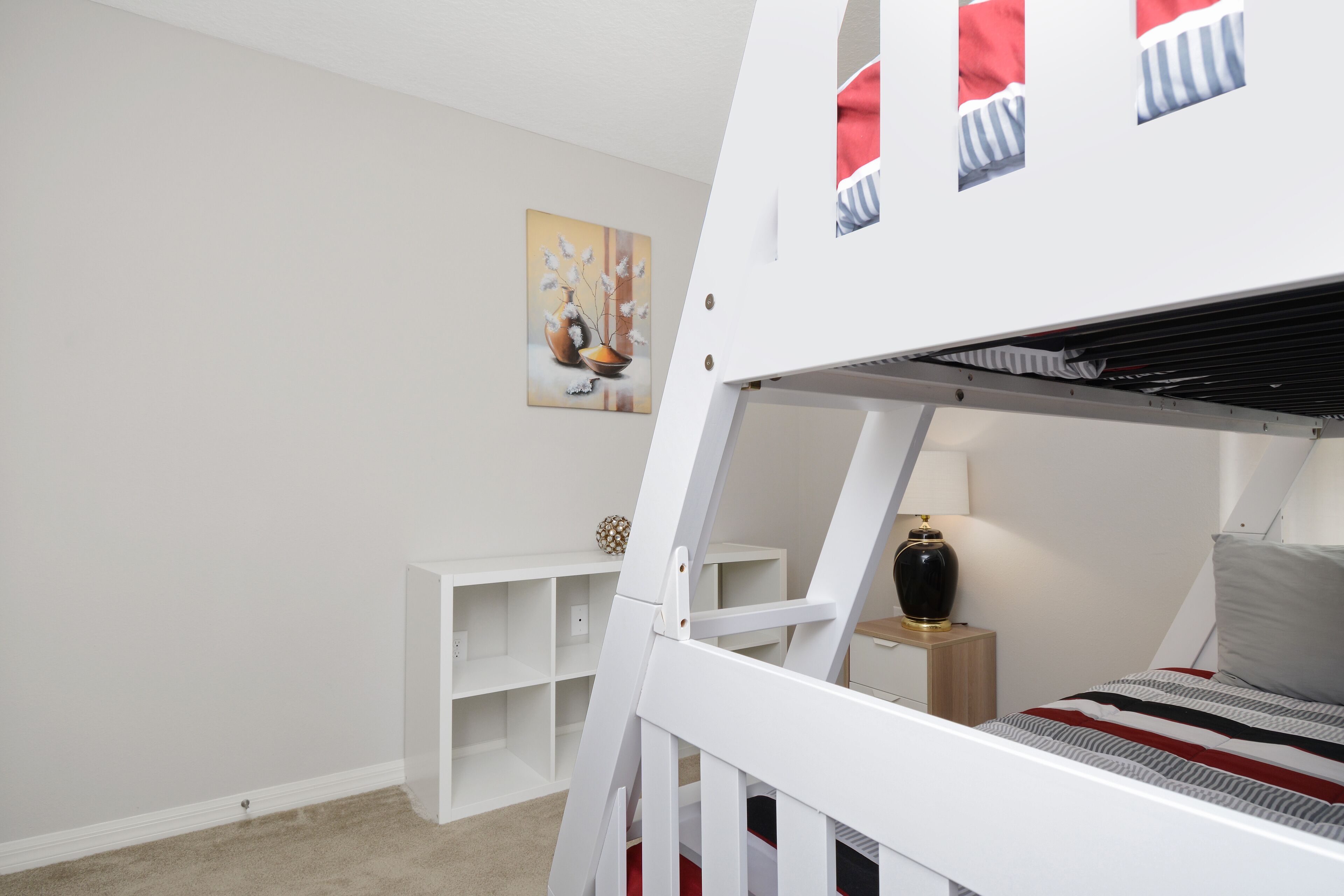 A bunk bed room with a white wall, a small shelf, a lamp, and a painting.