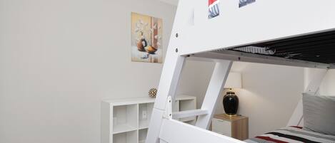 A bunk bed room with a white wall, a small shelf, a lamp, and a painting.