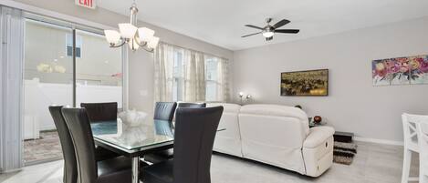 A modern living room with a glass dining table, black chairs, a white sofa, and a ceiling fan.