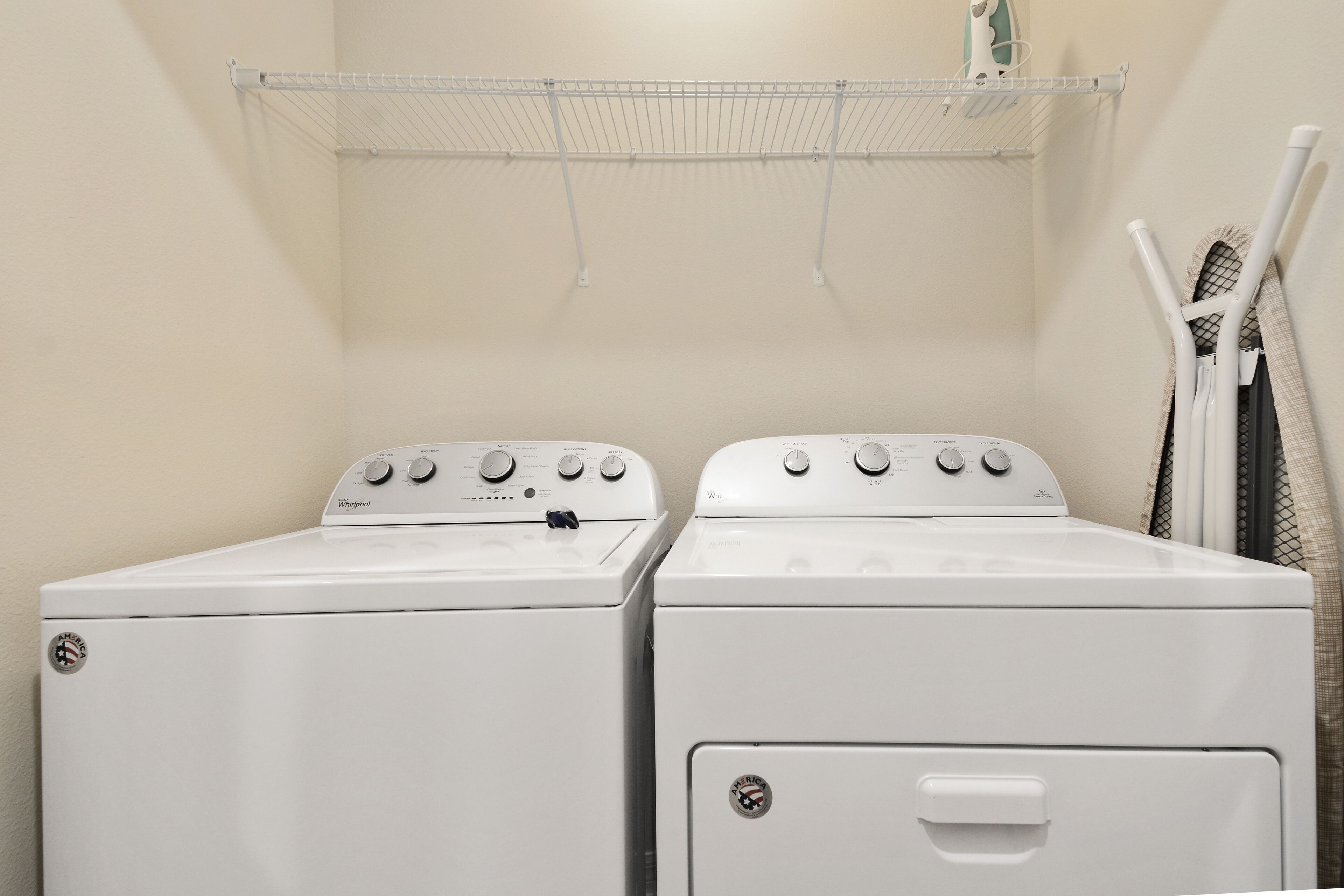 A laundry room with two stacked front-loading washers and dryers, a metal shelf above, and a drying rack to the right.