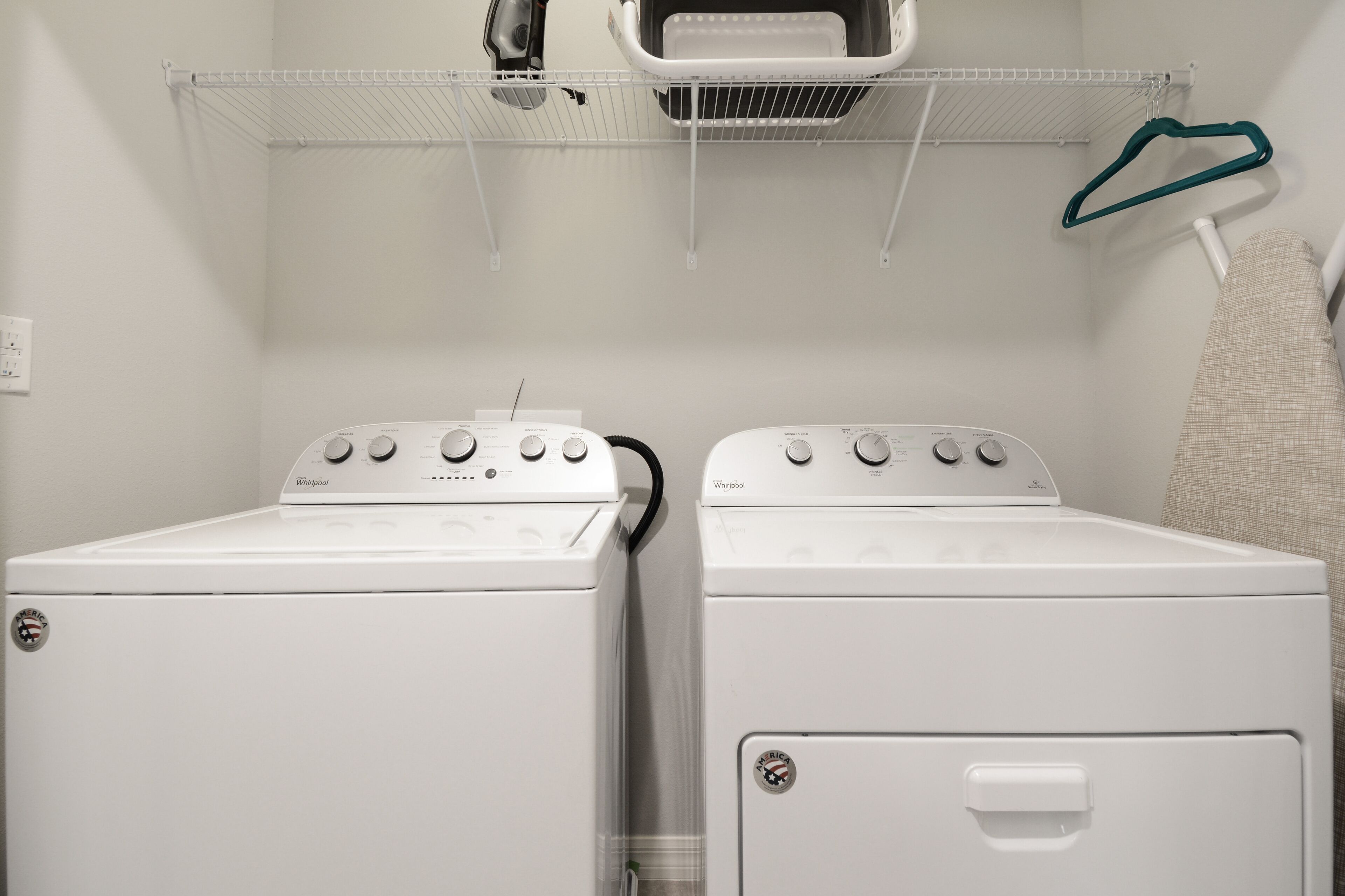 Egy mosókonyha, melyben két fehér Whirlpool márkájú szárítógép található, és a szárítógépek fölött egy fém polc van.
