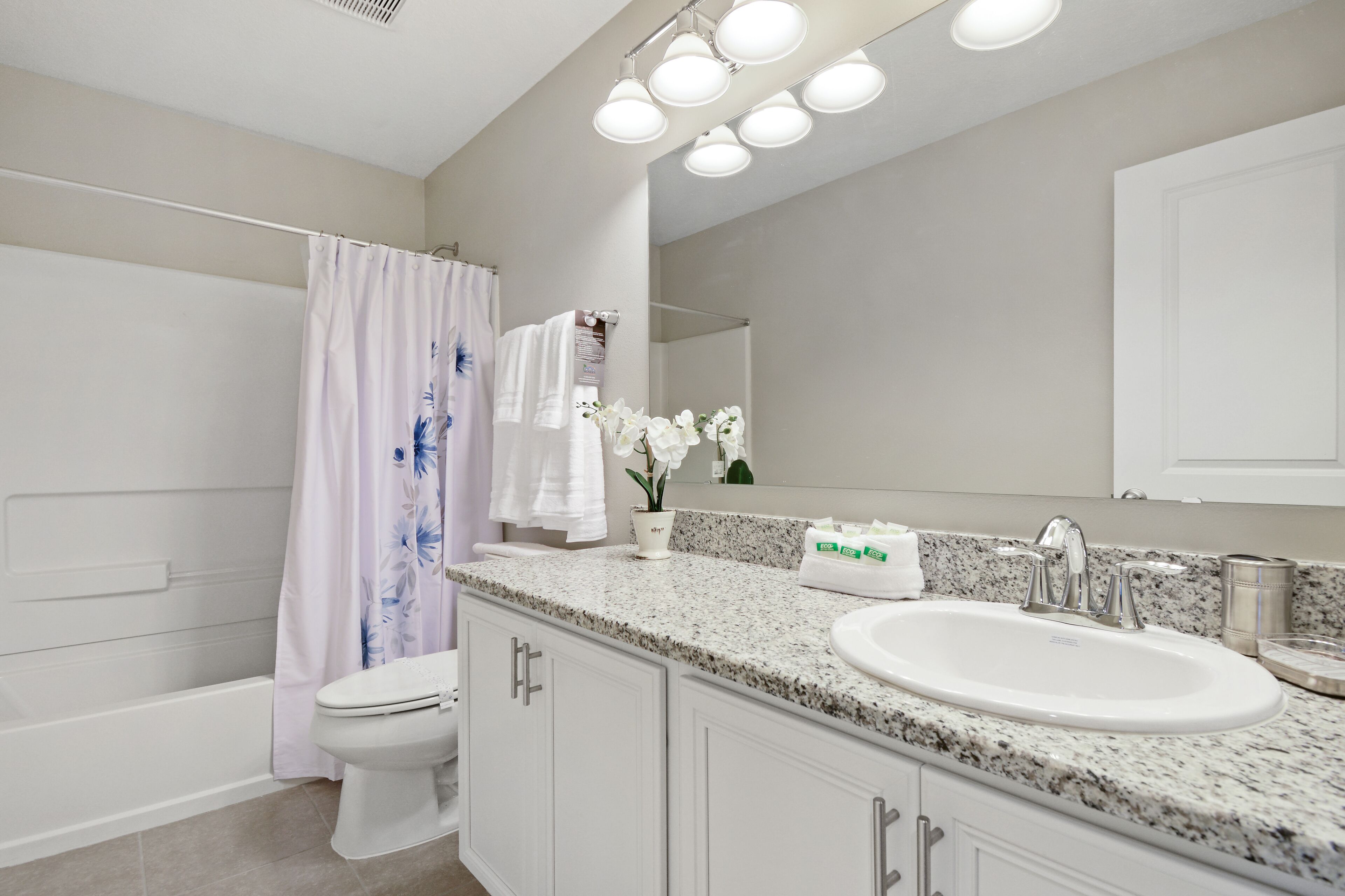 Une salle de bain moderne avec des toilettes blanches, une douche avec un rideau à motifs floraux, un lavabo sur un plan de travail en granit et un grand miroir au-dessus du lavabo.