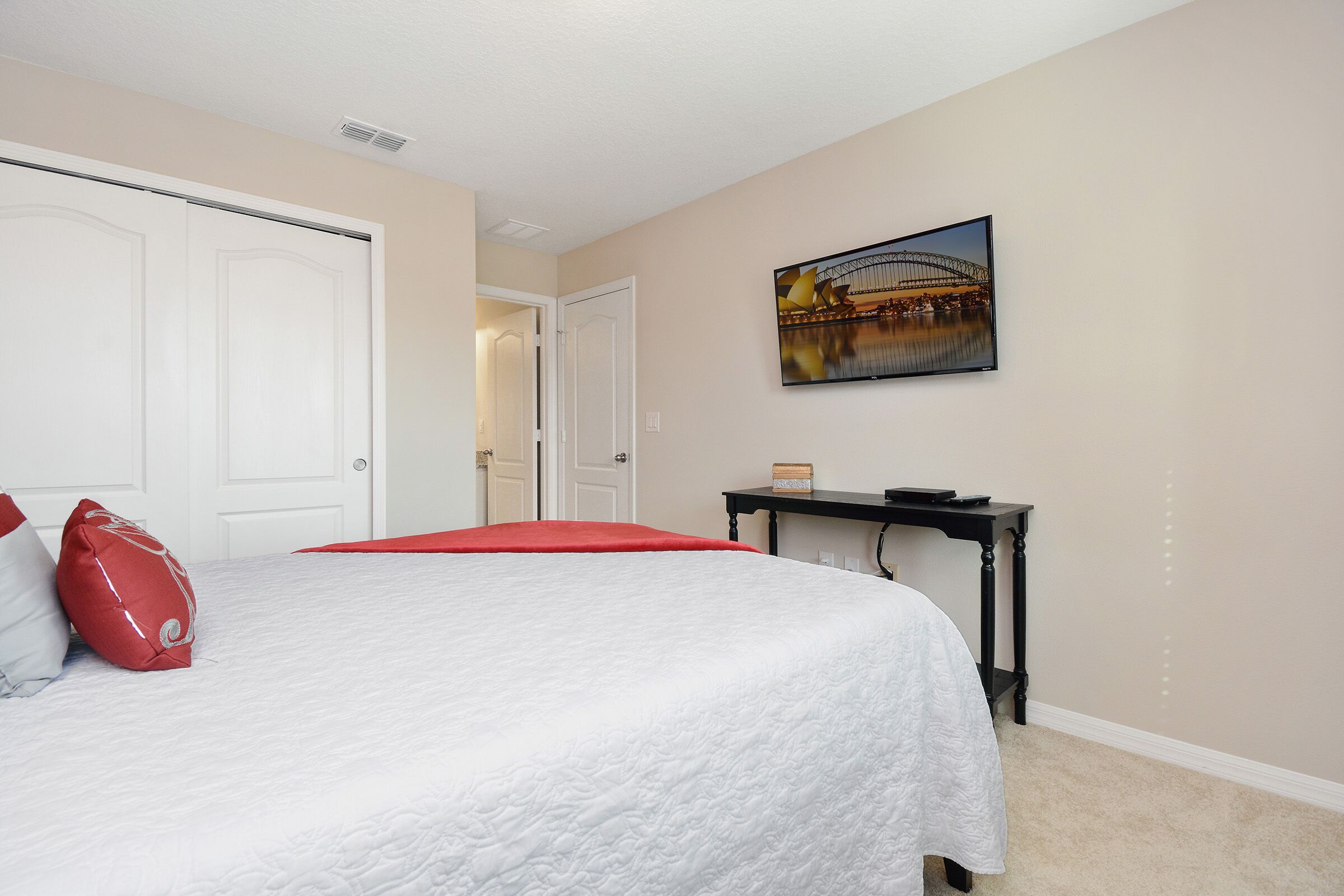 Une chambre à coucher avec un lit, un oreiller rouge, un oreiller blanc, un tableau représentant un pont et une petite table sur laquelle se trouvent un livre et une télécommande.