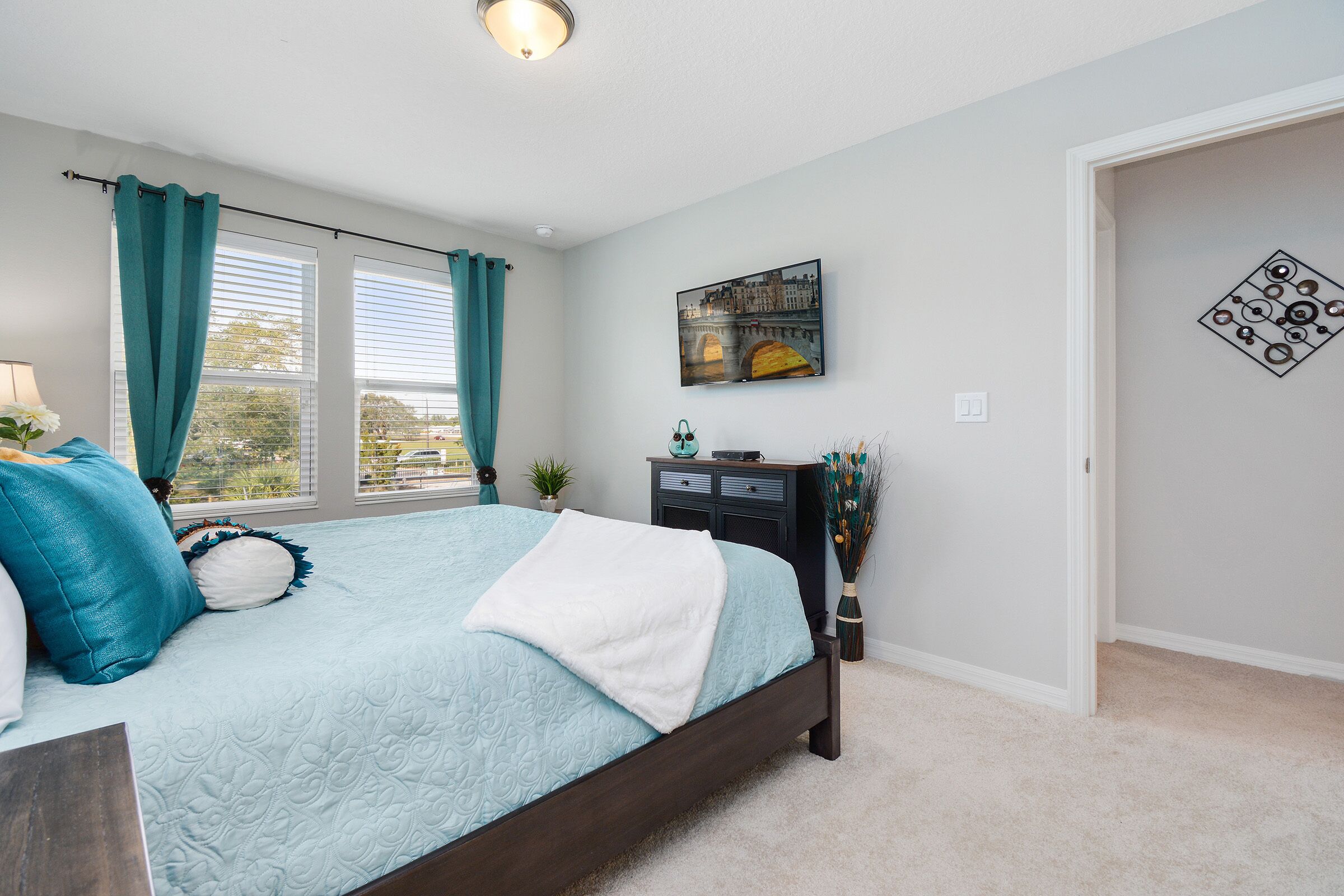 Une chambre à coucher comprenant un lit, une table de chevet, une commode, une fenêtre avec des rideaux, un tableau accroché au mur et une porte.