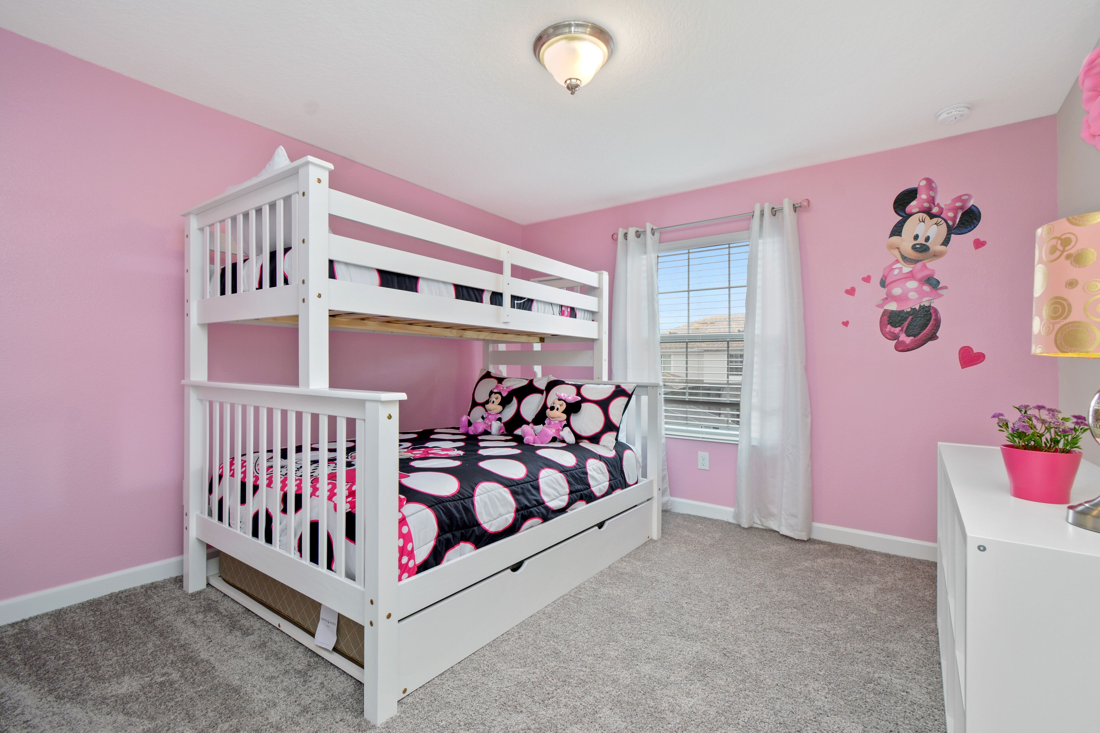 A bunk bed with a Minnie Mouse themed bedspread, a white desk with a pink flower pot, and a pink wall with a Minnie Mouse mural.
