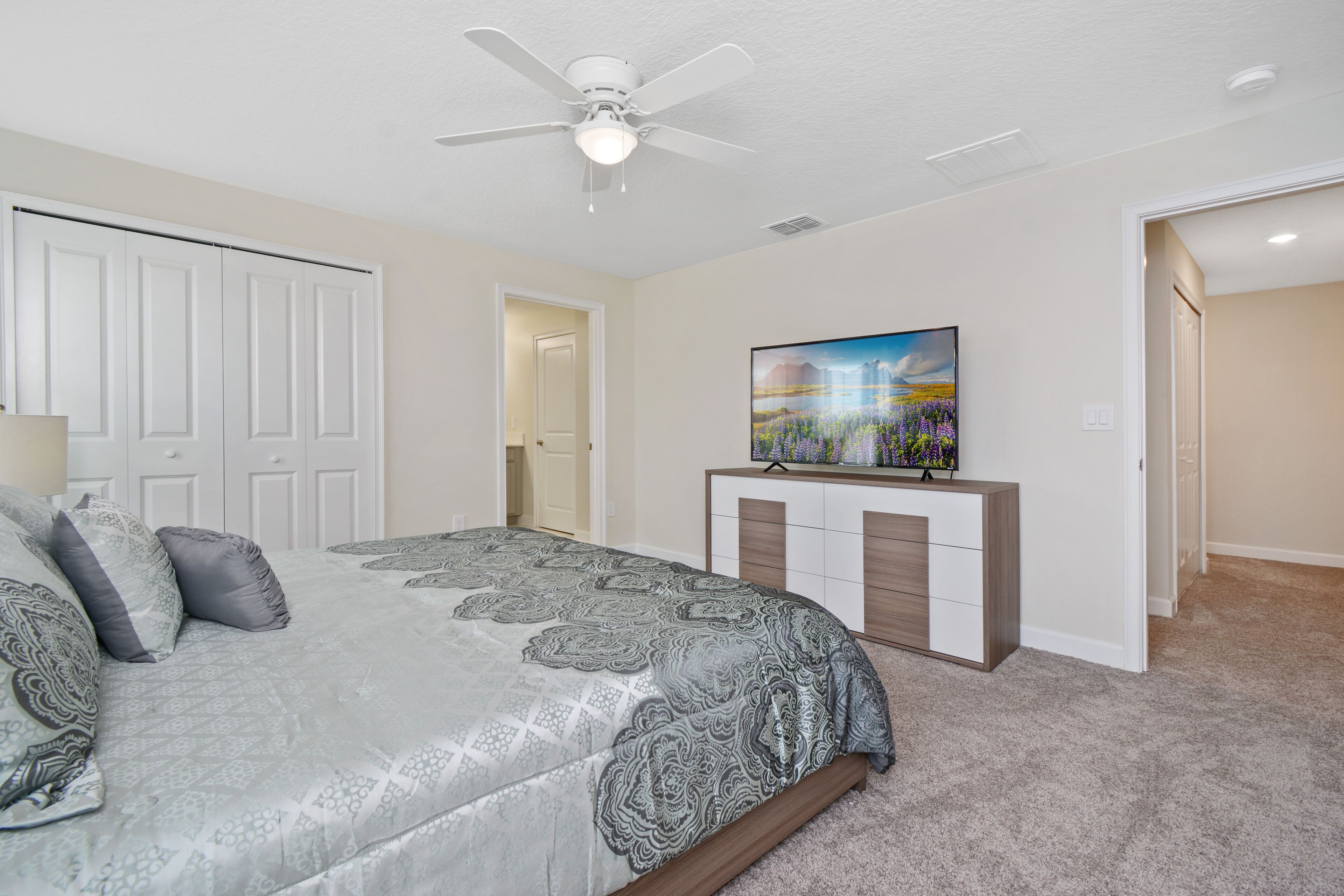 A bedroom with a bed, a ceiling fan, a TV on a stand, and a door leading to another room.
