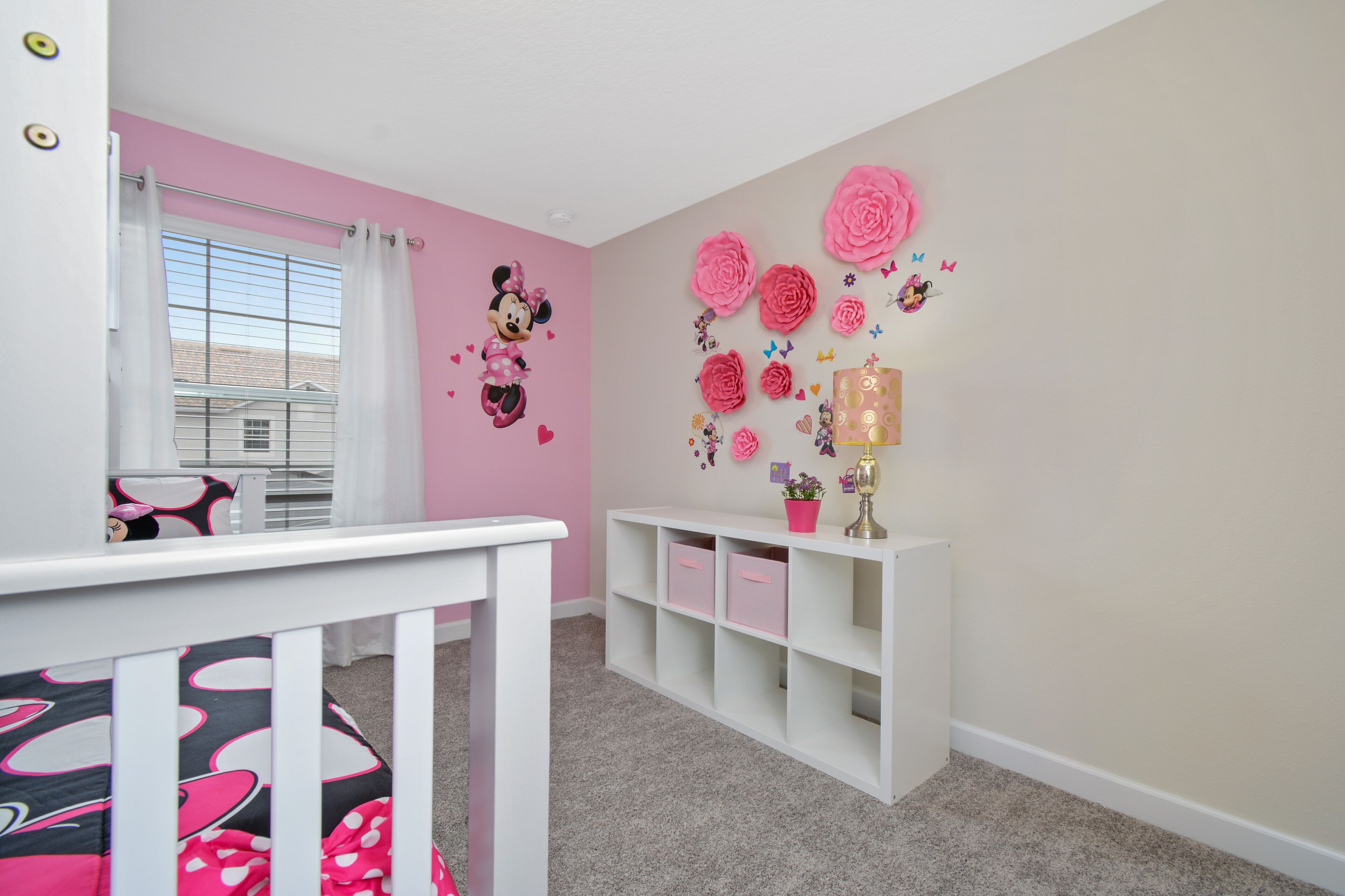 Ein Zimmer mit rosa und weißen Wänden, einem weißen Babybett, einem weißen Konsolentisch mit Stauraum und einem Fenster mit Blick auf Gebäude.