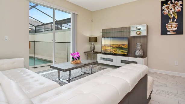 Una sala de estar moderna con un sofá, una mesa de centro y un televisor de pantalla plana sobre un mueble. Hay una puerta corrediza de vidrio que da a un balcón con piscina.