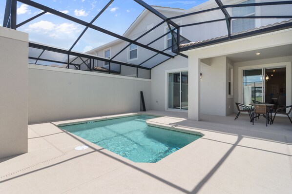 Una piscina con techo de vidrio, un patio con mesa y sillas, y una casa a lo lejos.
