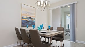A dining area with a table set for six, six chairs, a chandelier, and a large landscape painting on the wall. - Four Bedroom Close to Disney w/ Screened Pool @StoreyLake 4910 (Kissimmee)
