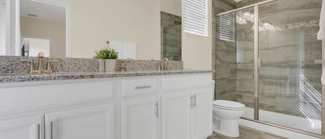 A modern bathroom with a granite countertop, white cabinetry, a glass shower enclosure, and a large mirror.