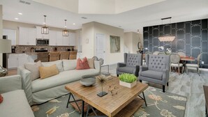 Ein geräumiger Wohnbereich mit Sofa, Sesseln und einem Esstisch. Im Hintergrund ist eine Küche mit weißen Schränken und einer Mikrowelle zu sehen.
