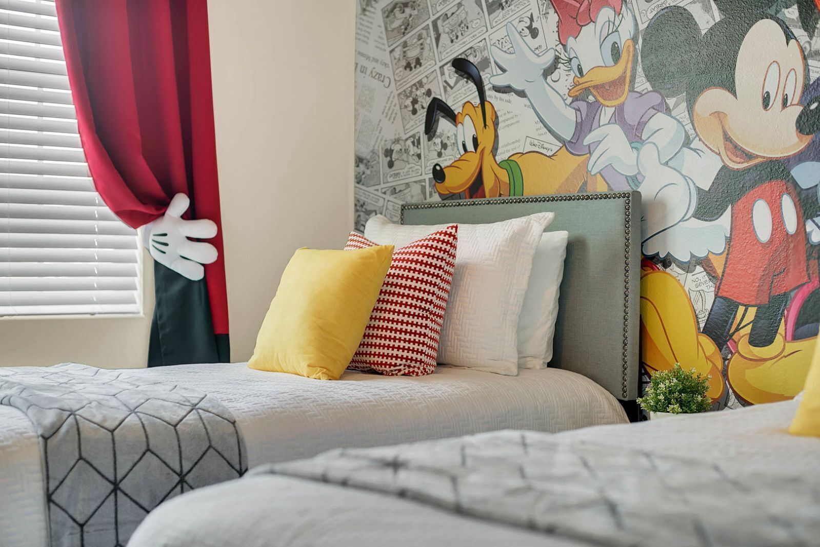 A hotel room with two beds, a Mickey Mouse mural on the wall, and a window with red curtains.