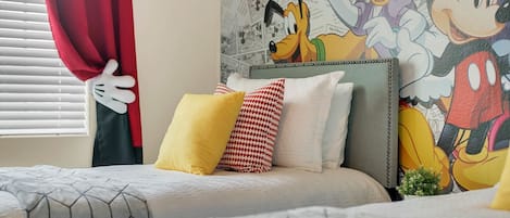 A hotel room with two beds, a Mickey Mouse mural on the wall, and a window with red curtains.