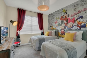 A hotel room with two beds, red curtains, a mural with cartoon characters, a desk with a lamp, and a view of a mountain through the window.
