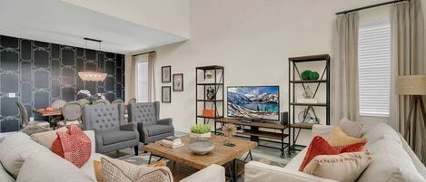 A spacious living room with a high ceiling, a large flat-screen TV, and a dining area with chairs and a table.