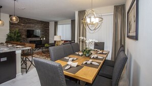 A modern dining area with a wooden table set for six, surrounded by upholstered chairs. A large chandelier hangs above the table, and a television is mounted on the wall to the left.