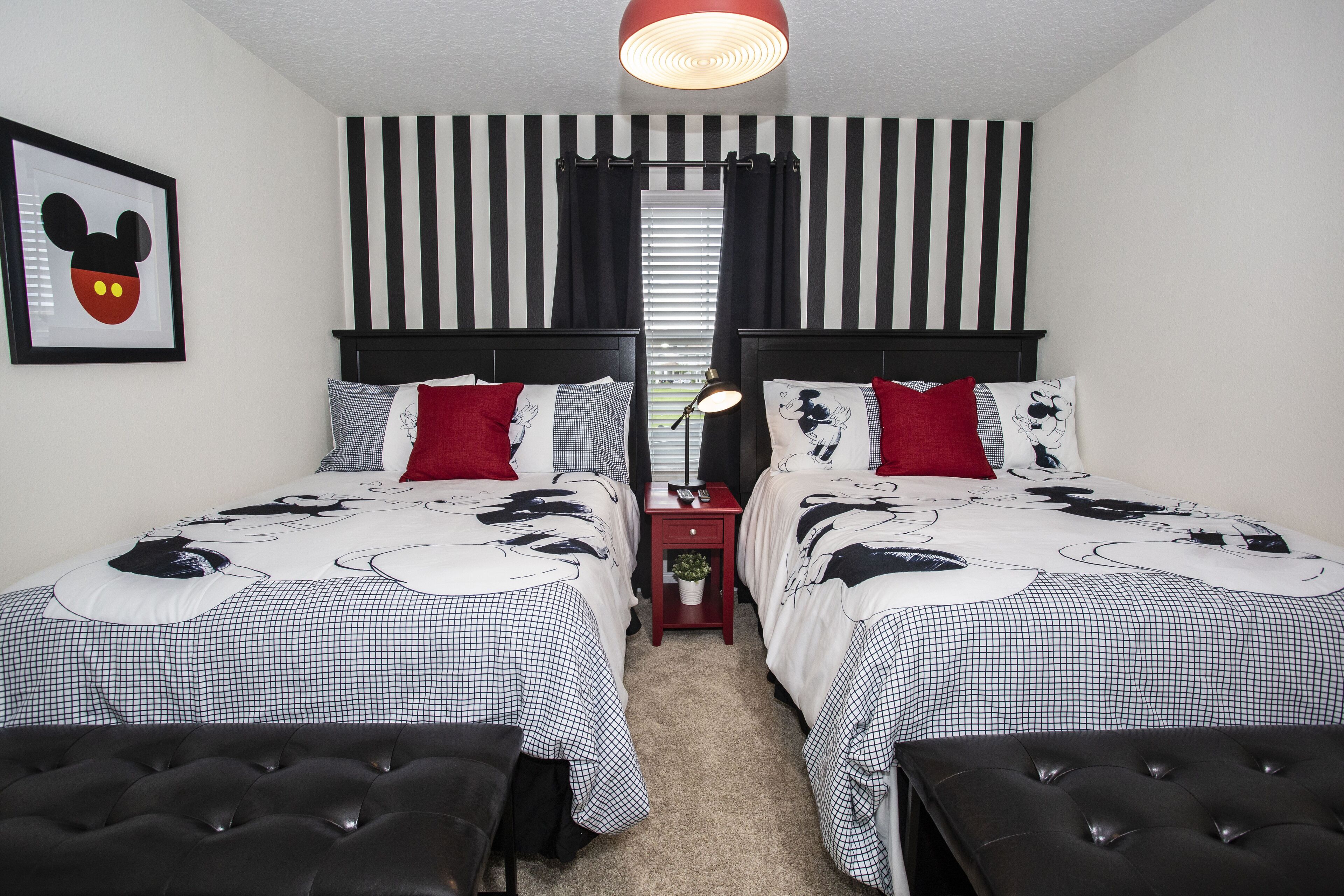 A bedroom with two beds, striped curtains, Mickey Mouse themed bedding, and a red nightstand with a lamp.