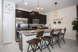 A modern kitchen with dark cabinets, stainless steel appliances, a large island with bar stools, and a wall clock. - Four Bedroom @ Storey Lake Resort Close to Disney w/ Pool 3063 (Kissimmee)