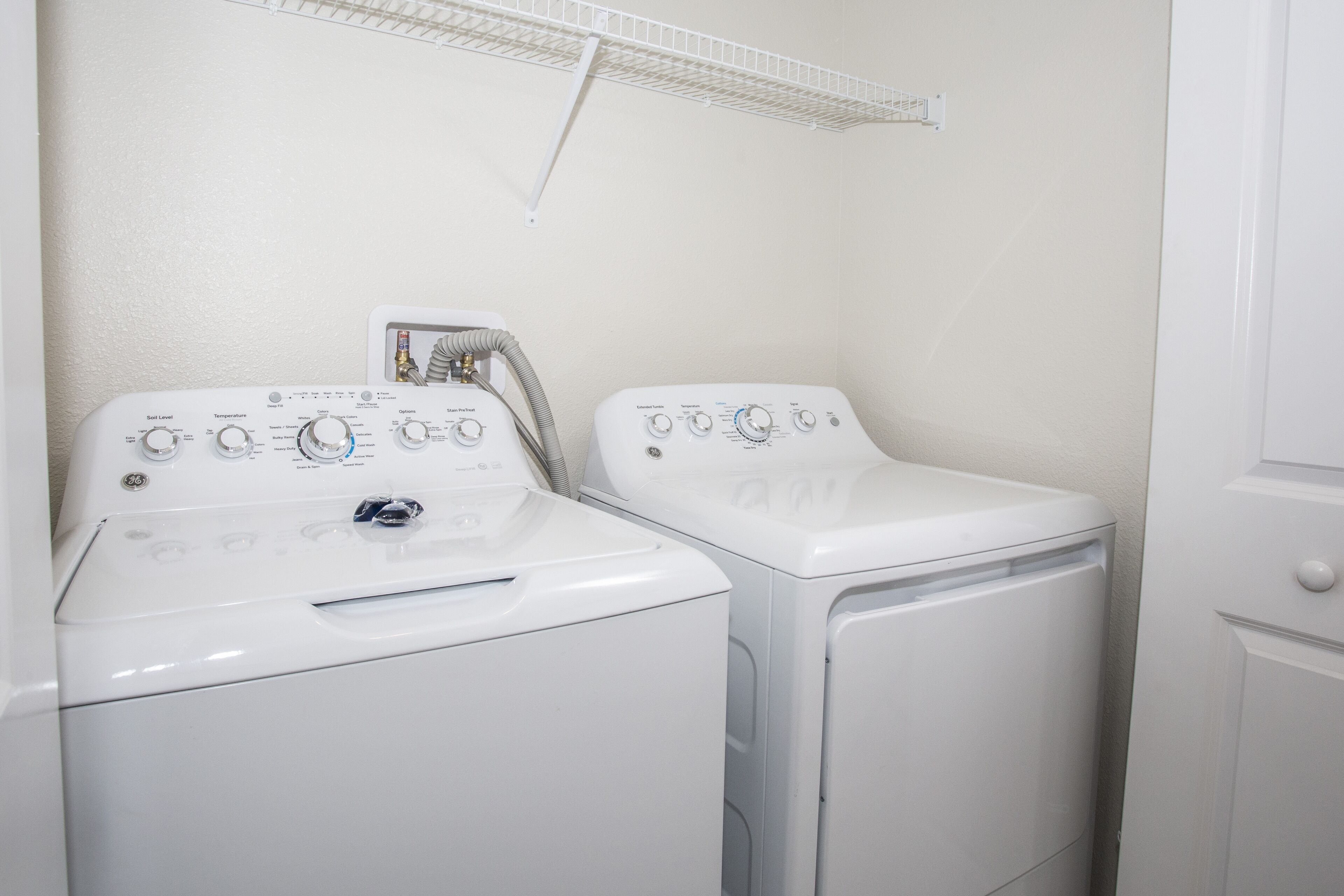 Two front-loading washing machines in a laundry room with a white wall and a metal shelf above.