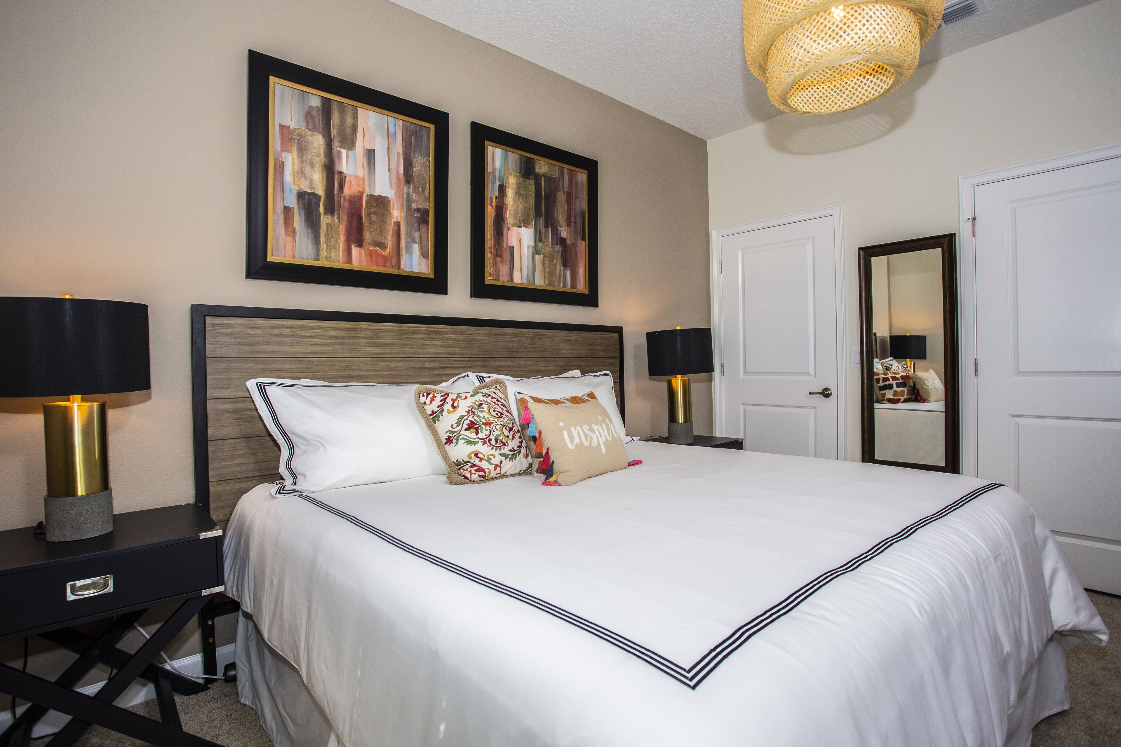 A neatly made bed with white linens and decorative pillows, a bedside table with a lamp, and two framed abstract paintings on the wall.