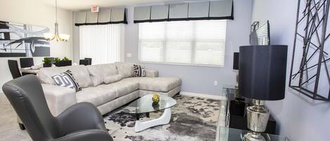 Una sala de estar moderna con un sofá seccional, una mesa de centro de vidrio, un sillón negro y una lámpara de pie.