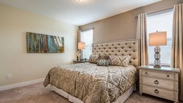 A bedroom with a tufted headboard, a large bed with patterned bedding, two bedside tables with lamps, a wall-mounted abstract painting, and a window with blinds.