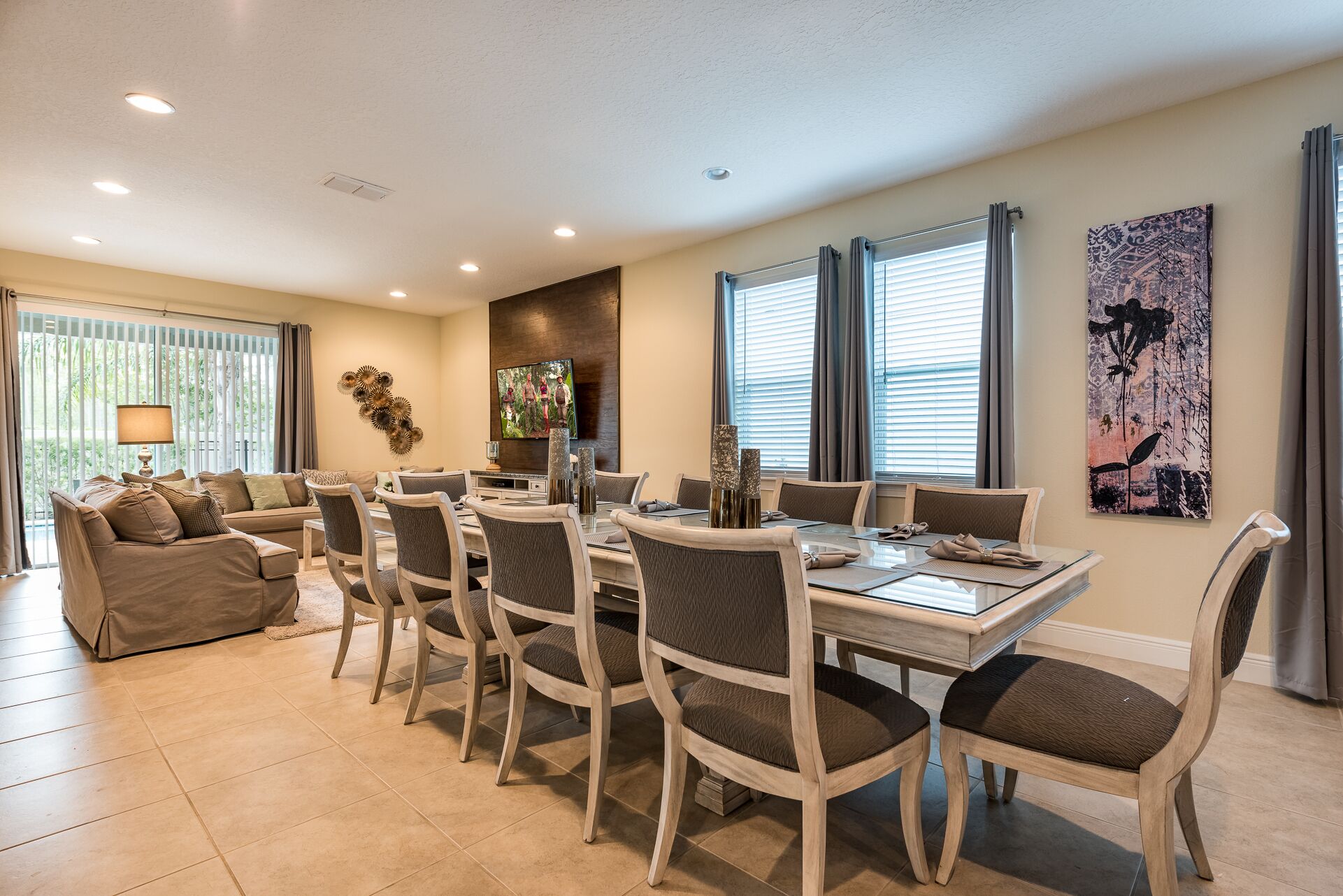 A spacious dining area with a large table and multiple chairs, a sofa, and a television mounted on the wall.