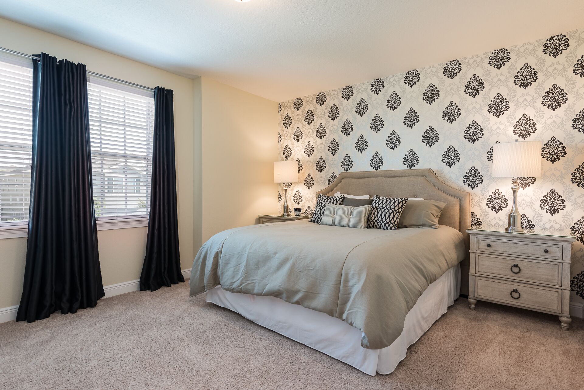 A bedroom with a bed, two bedside tables, a nightstand, a window with curtains, and patterned wallpaper.