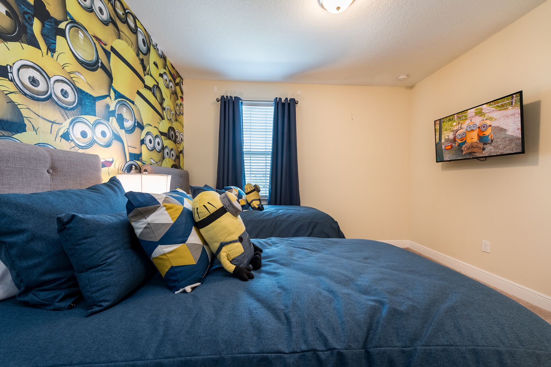 A bedroom with two beds, a Minion-themed wall, a window with curtains, and a painting on the wall.