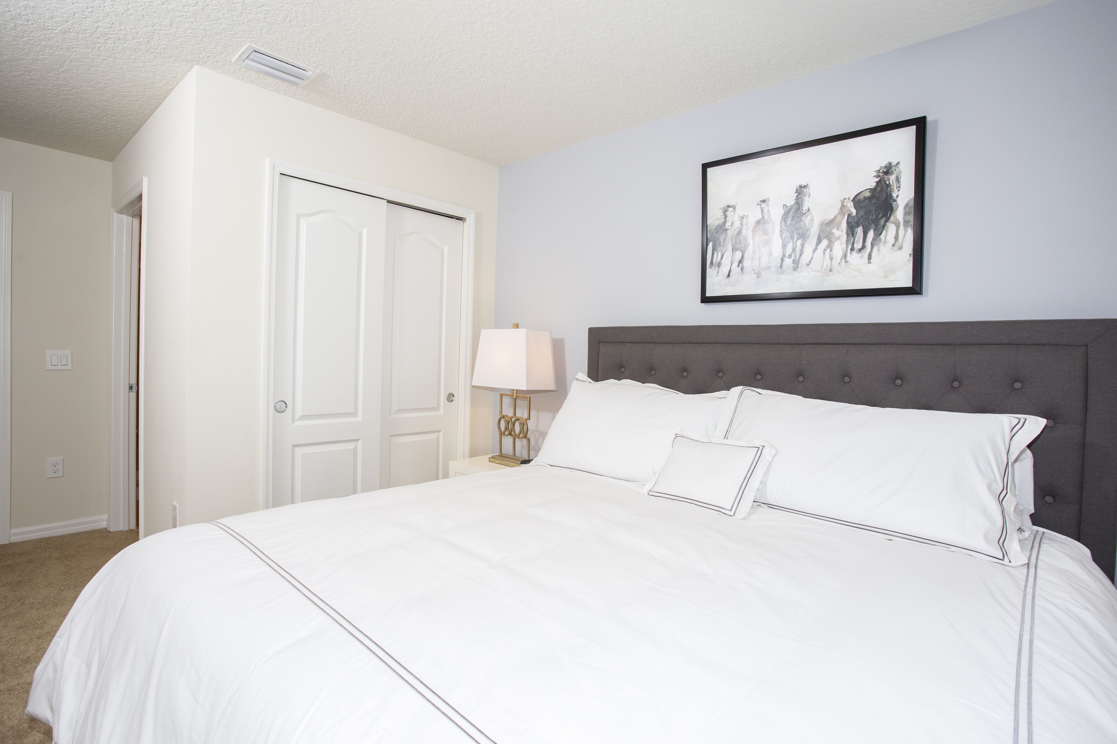 A bedroom with a large bed, a grey headboard, a bedside table with a lamp, and a framed picture on the wall.