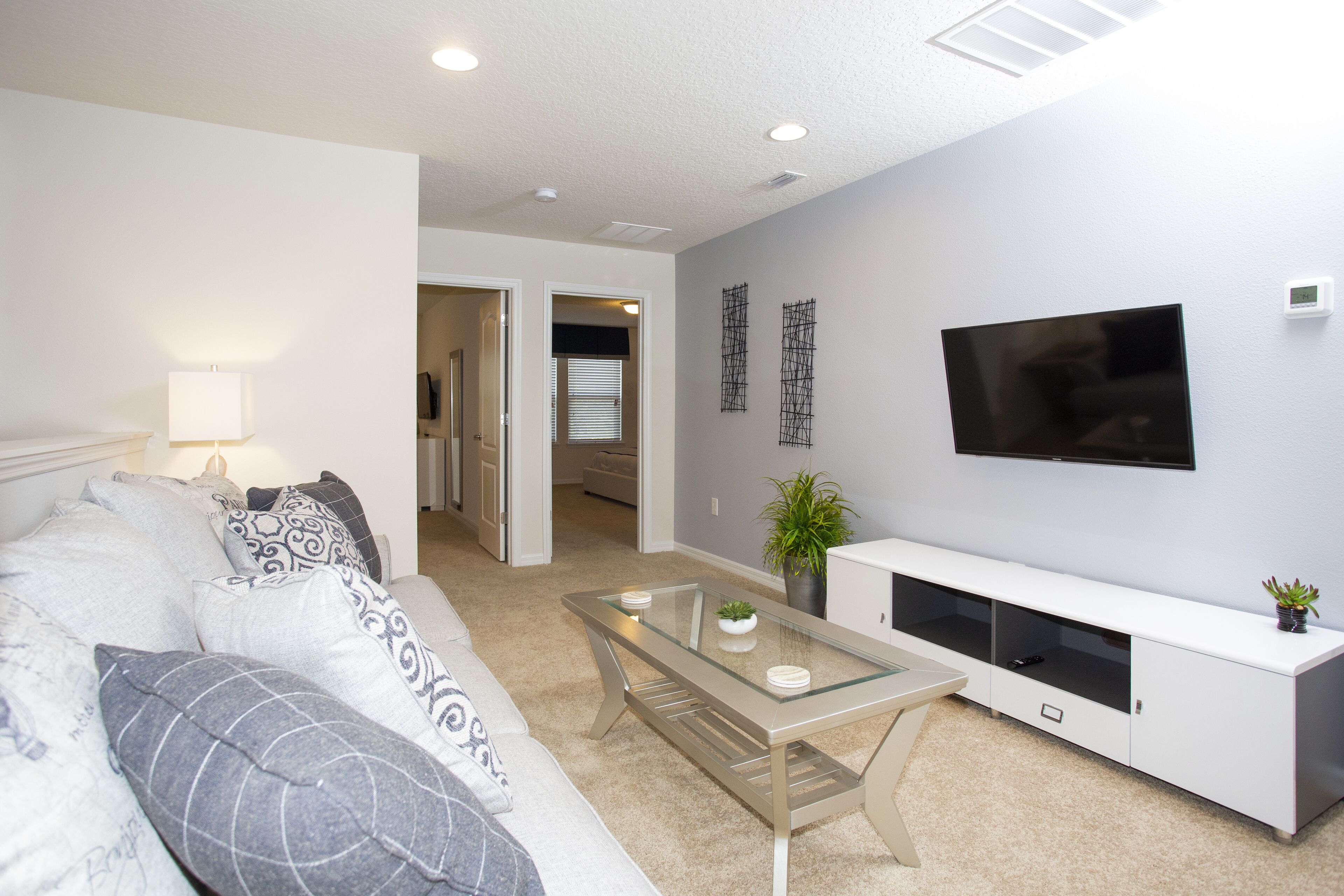 A modern living room with a sofa, a glass-top coffee table, a flat-screen TV mounted on the wall, and a white entertainment unit.