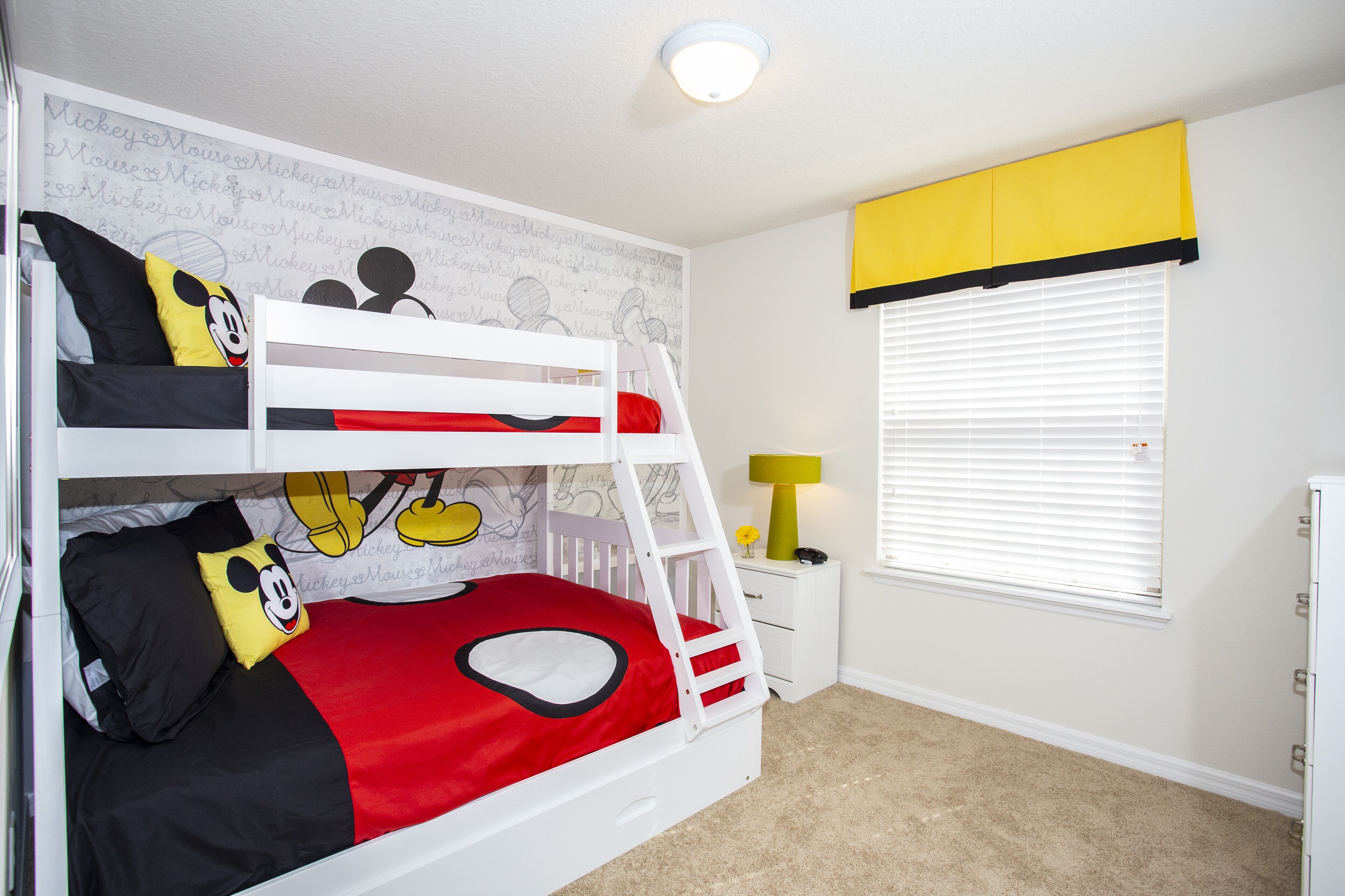 Un lit superposé avec une parure de lit aux motifs de Mickey Mouse, une petite table de chevet avec une lampe et une fenêtre avec des stores.