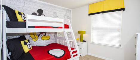 A bunk bed with Mickey Mouse-themed bedding, a small bedside table with a lamp, and a window with blinds.