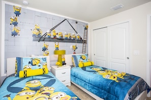 A bedroom with two beds, each covered with a blue and yellow Minion-themed duvet. There is a white nightstand with a lamp and a shelf above the beds.