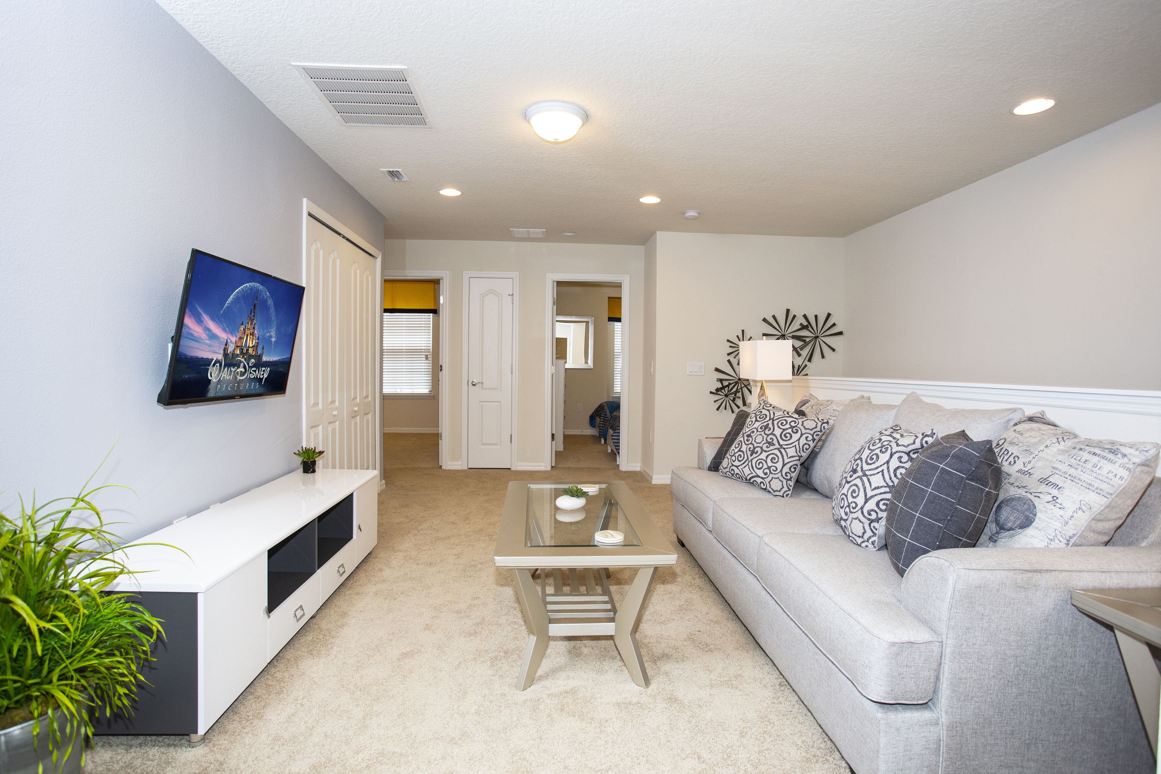 A living room with a flat-screen TV, a glass coffee table, a grey sofa with patterned cushions, and a potted plant.