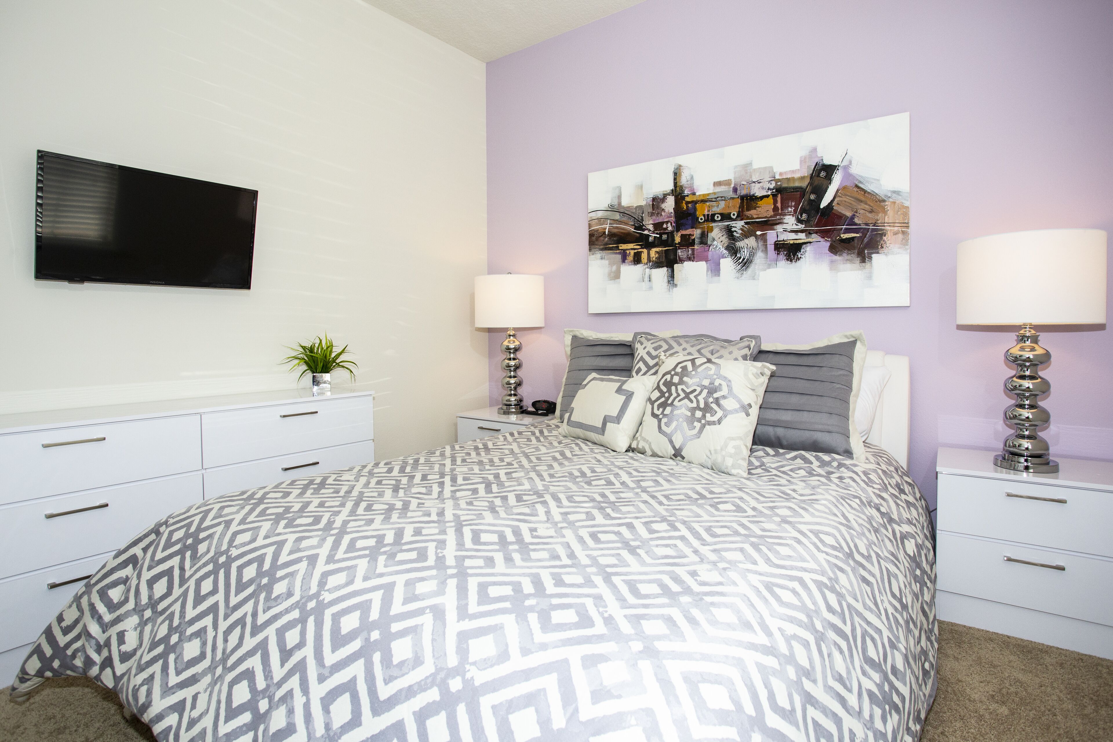 A bedroom with a patterned bedspread, a flat-screen TV, two bedside lamps, a nightstand with a plant, and a painting on the wall.