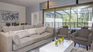 Un salon moderne avec un canapé gris, deux fauteuils, une table basse blanche et une grande fenêtre donnant sur l’extérieur.
