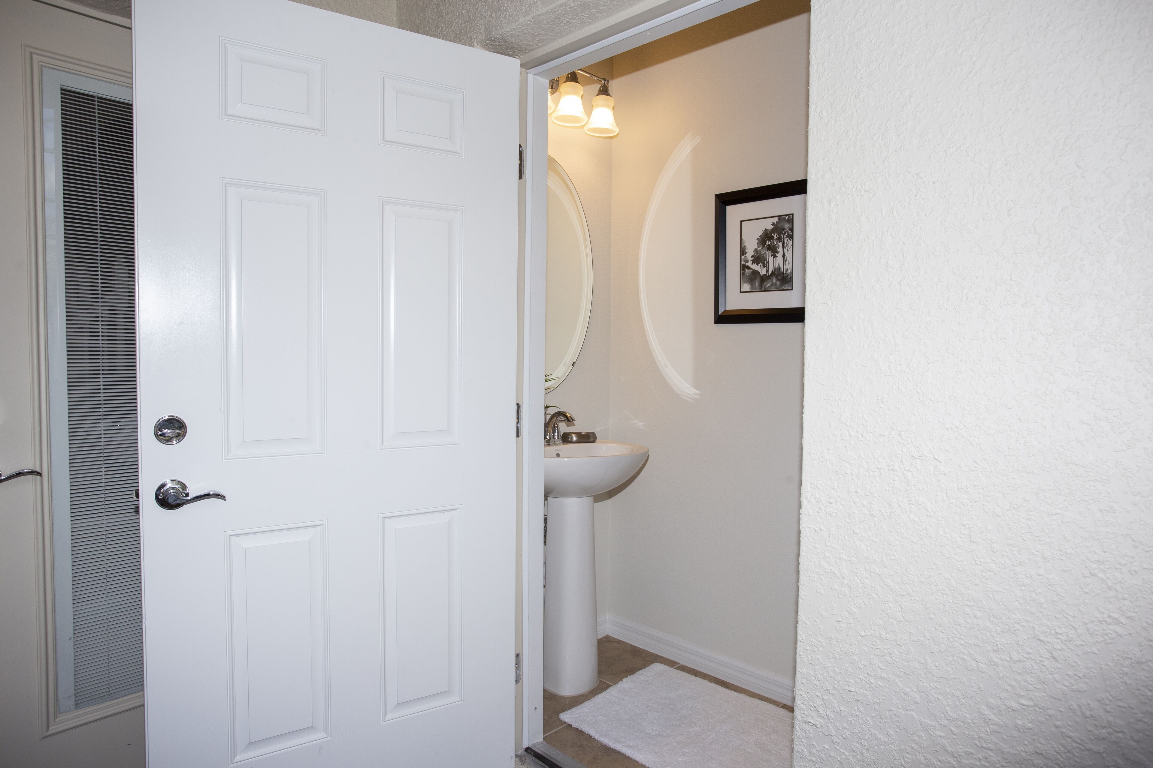 A white door with a silver handle, a white pedestal sink, a white towel on the floor, and a framed picture on the wall.