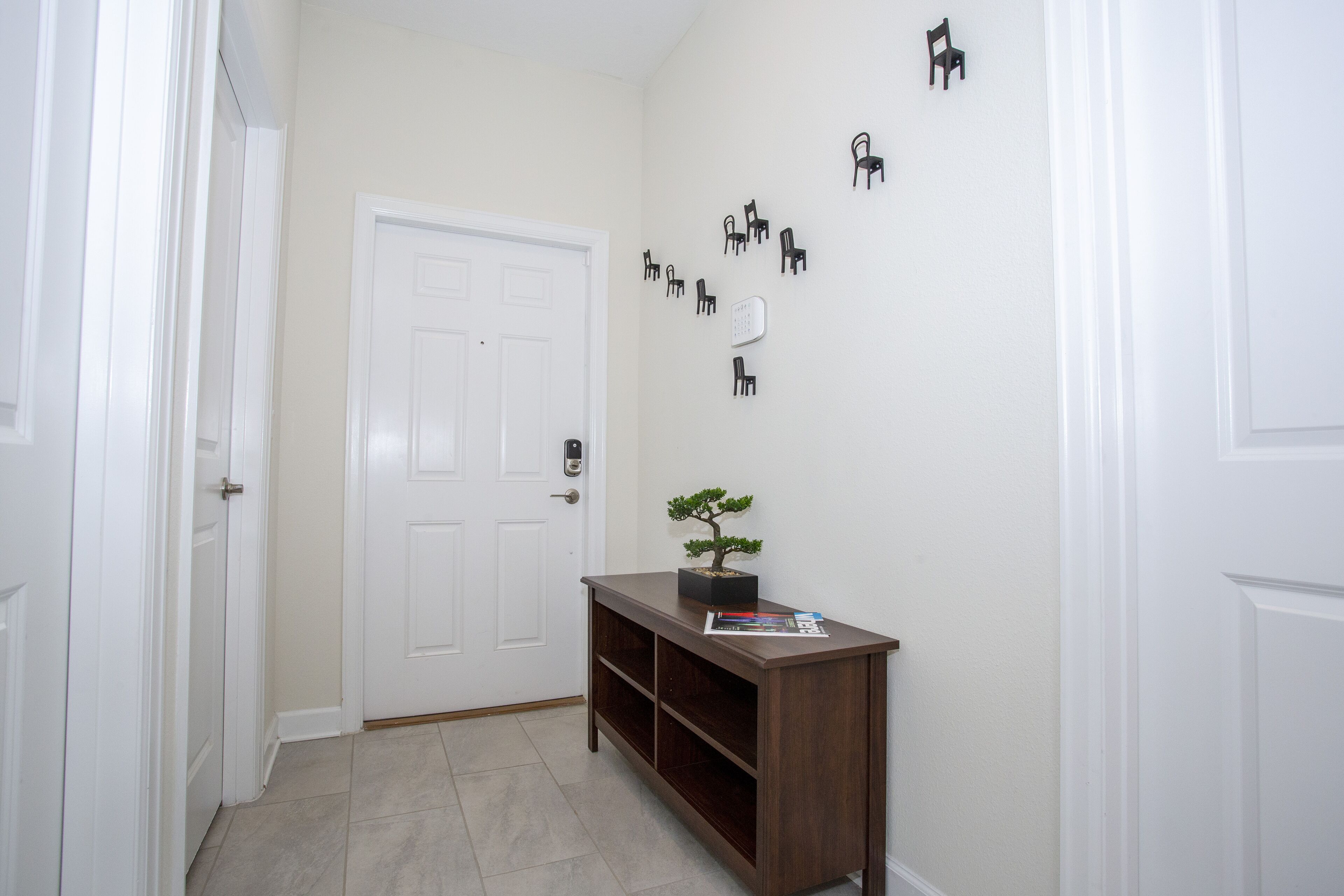 Un pasillo con una puerta blanca, una consola de madera con un bonsái y ganchos para abrigos en la pared.