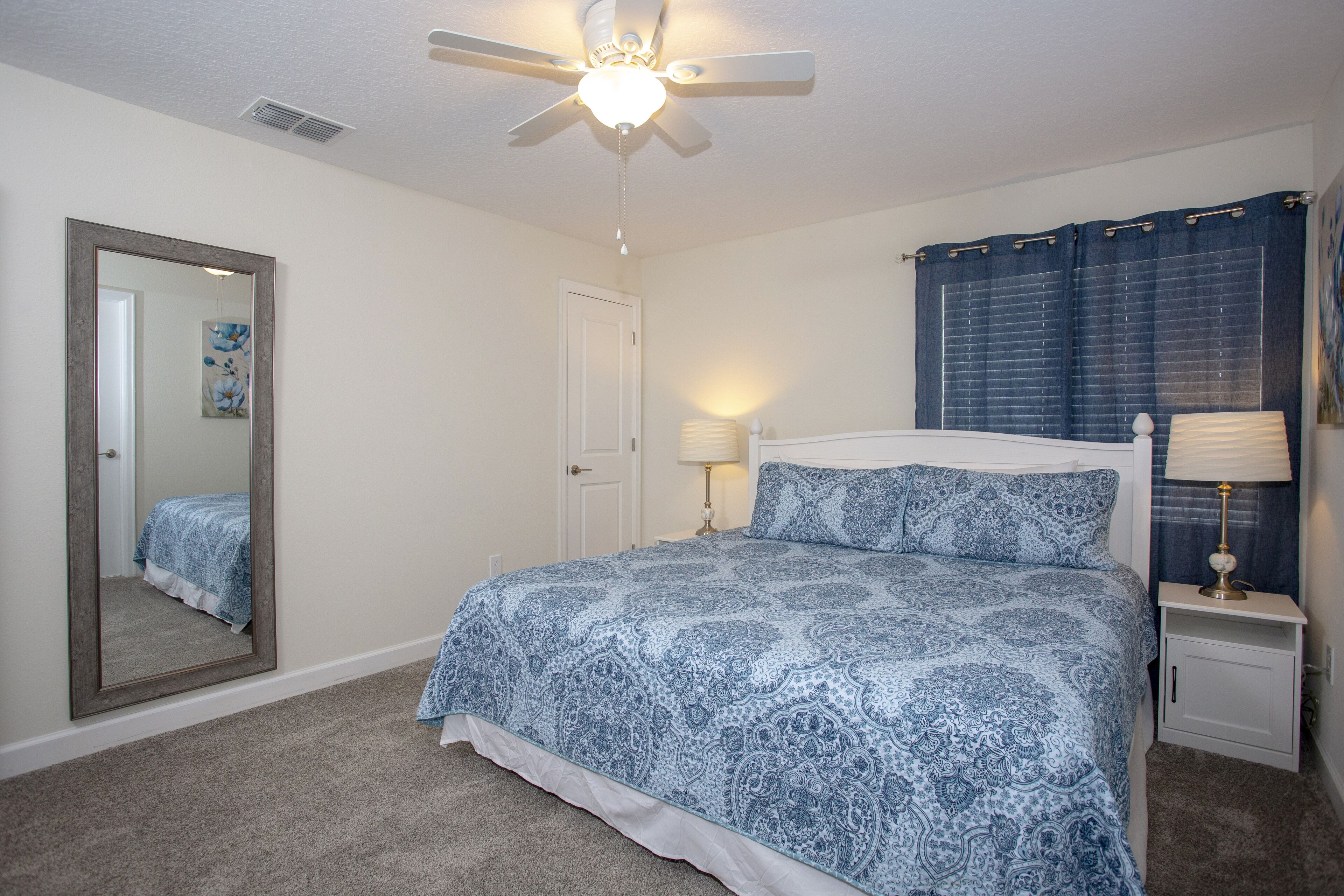 A bedroom with a bed, a ceiling fan, a nightstand with a lamp, a mirror, and a door.