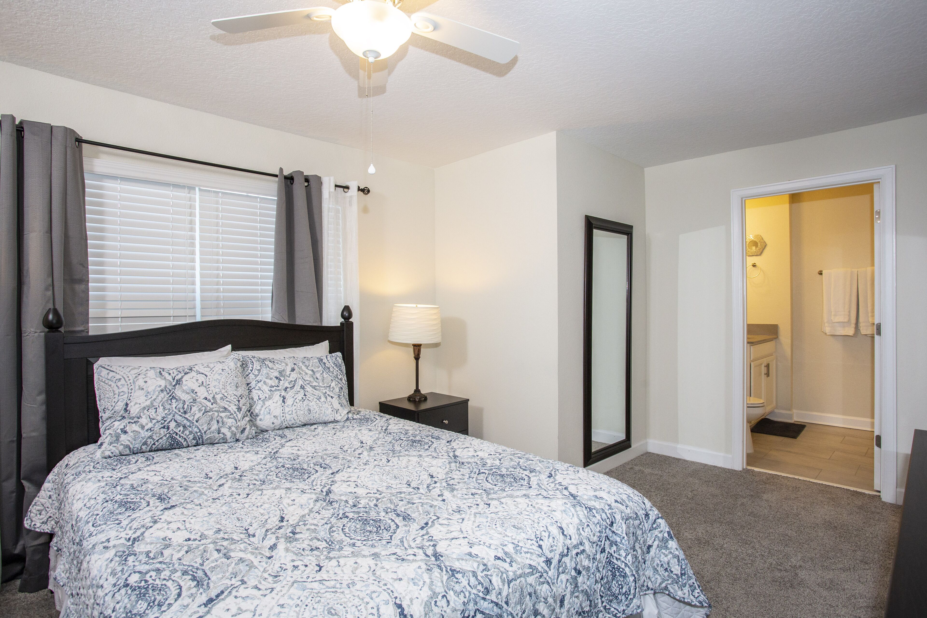 A bedroom with a bed, a nightstand, a lamp, a ceiling fan, a window with curtains, and a door leading to a bathroom.
