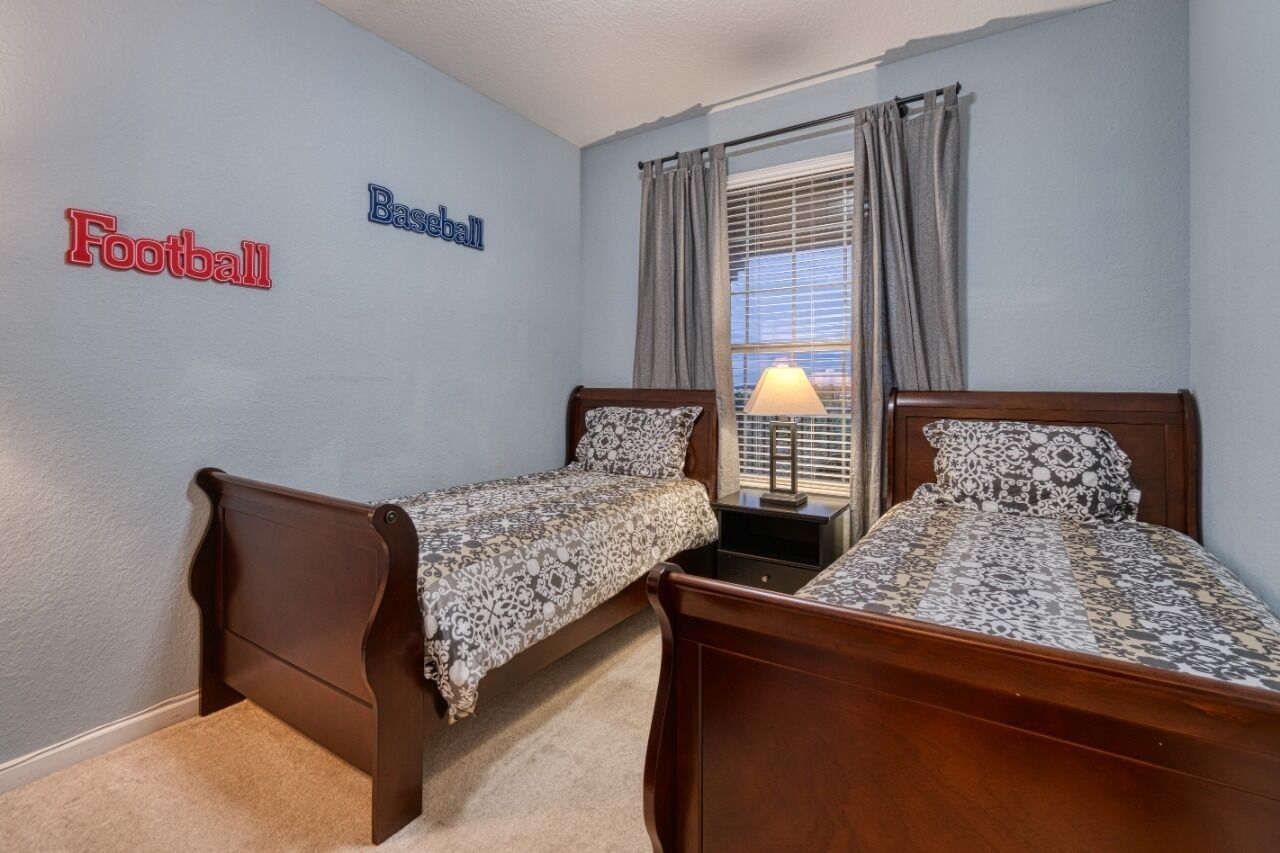 A bedroom with two single beds, a nightstand with a lamp, a window with curtains, and wall decorations labeled "Football" and "Baseball".