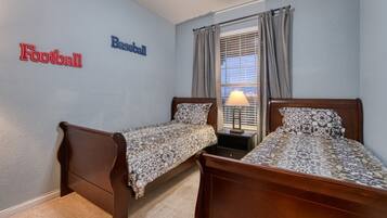 A bedroom with two single beds, a nightstand with a lamp, a window with curtains, and wall decorations labeled "Football" and "Baseball".