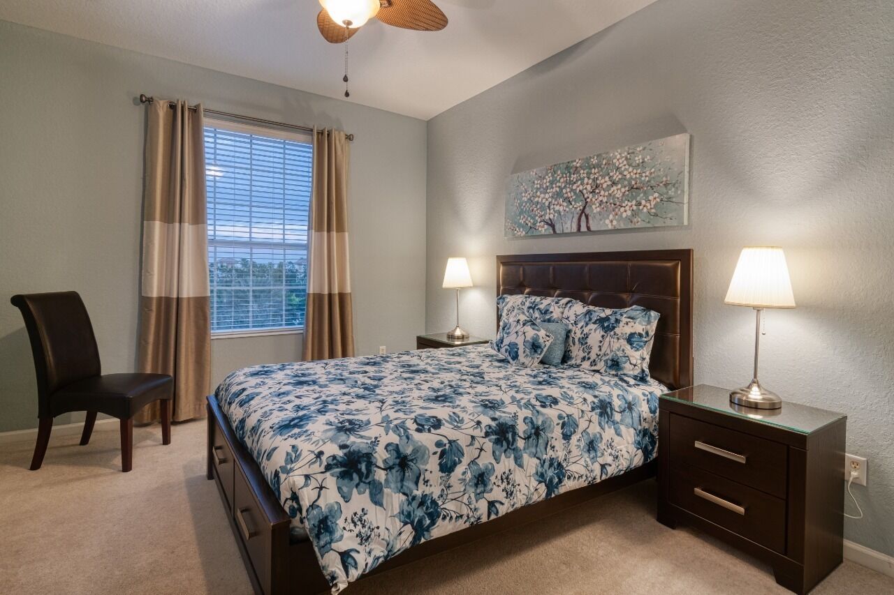 Un dormitorio con cama, dos mesitas de noche con lámparas, una silla, una ventana con cortinas y un cuadro en la pared.