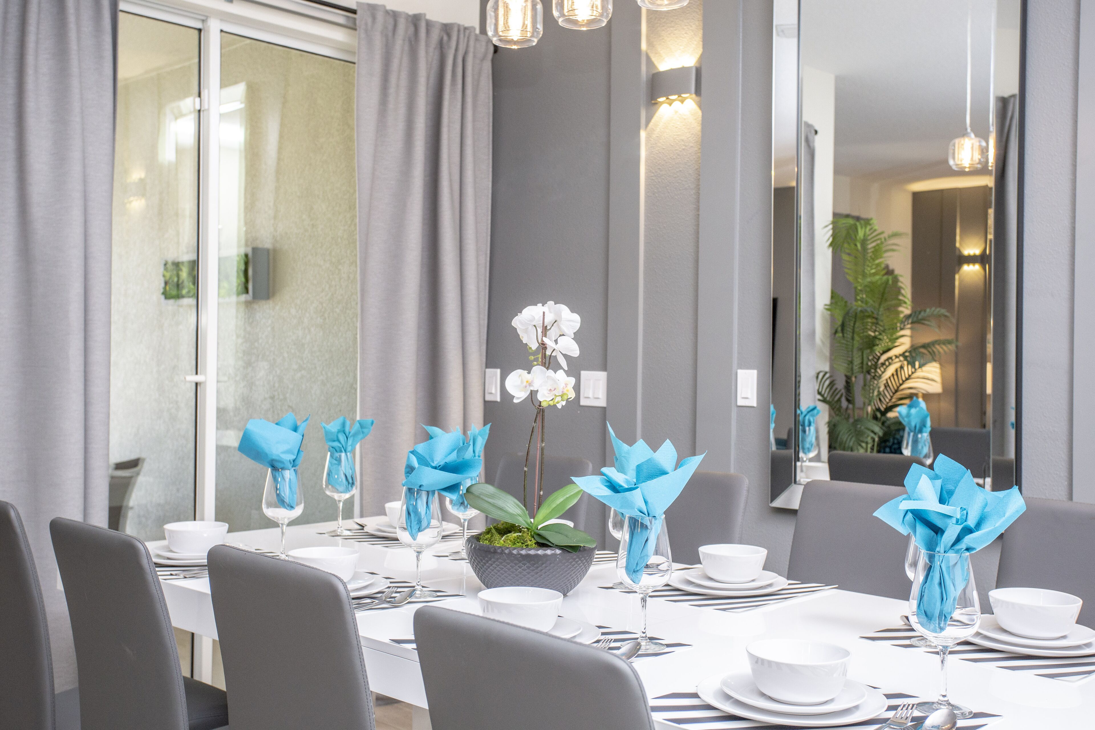 A modern dining area with a long table set for a meal, featuring white plates, silverware, and blue napkin decorations.