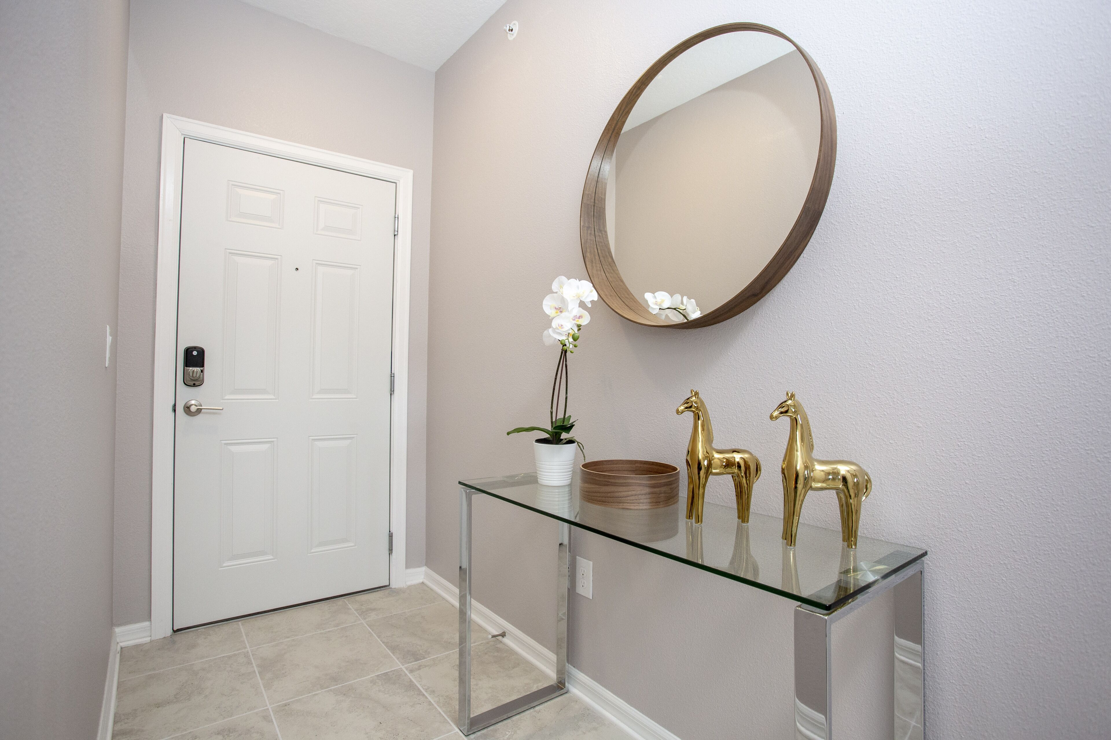 A hallway with a white door, a glass console table with a mirror, and two golden horse statues.