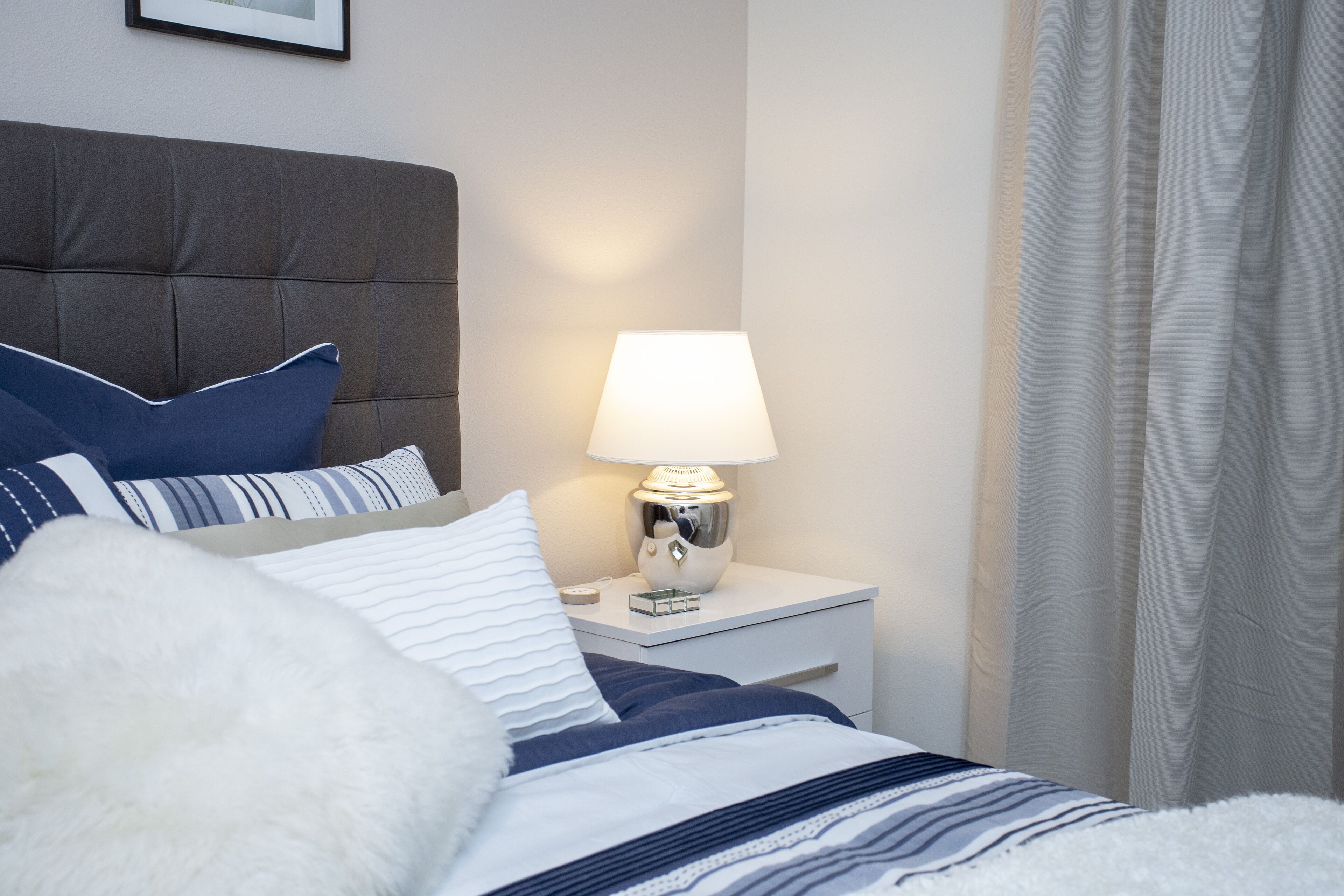 A neatly made bed with a striped duvet, a white fluffy pillow, and a blue pillow. There is a white bedside table with a lamp and a small clock. The room has a neutral color scheme with a grey headboard and white walls.