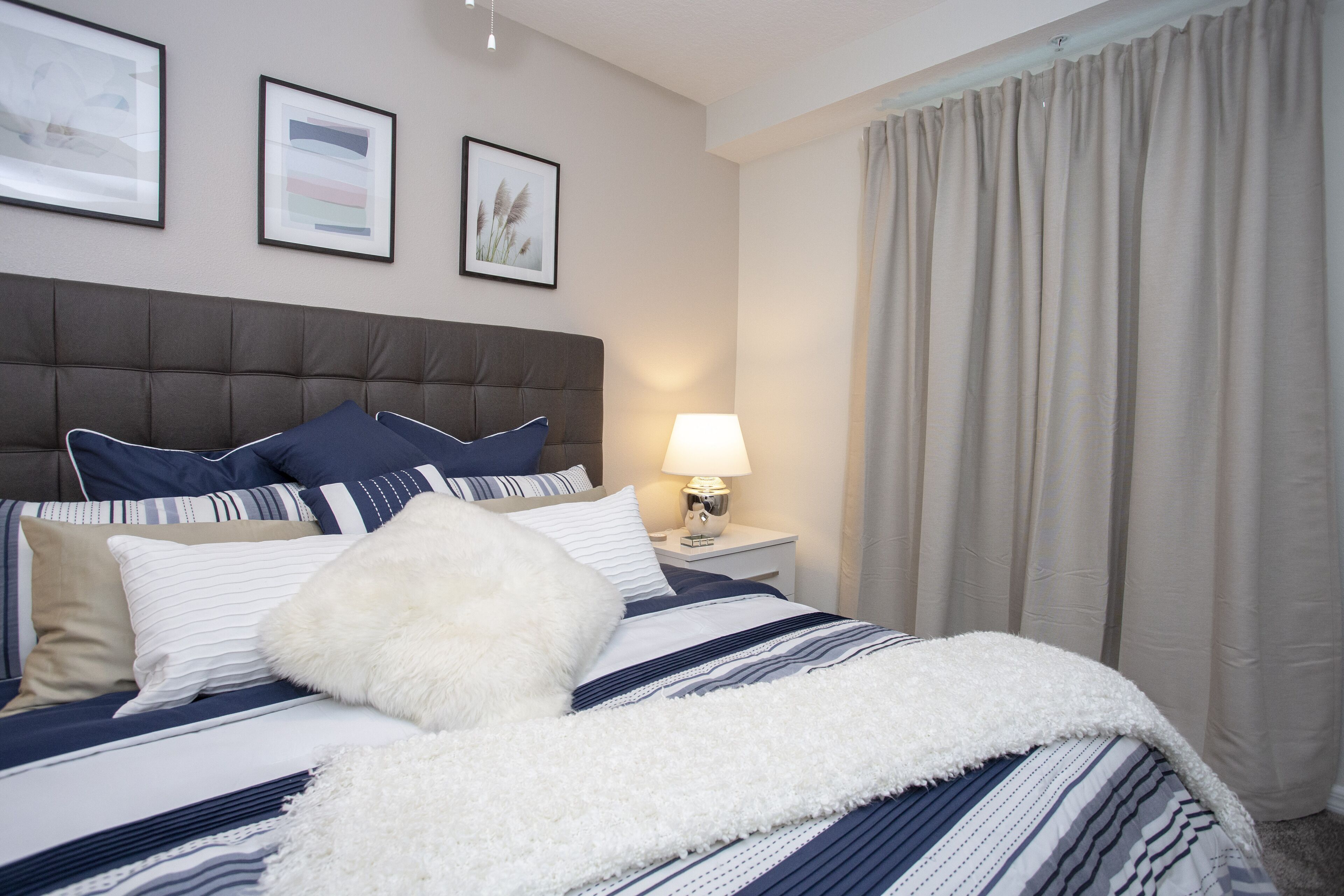 A neatly made bed with a striped bedspread, a fluffy white throw, and several pillows. There is a nightstand with a lamp and a framed picture on the wall above the bed.